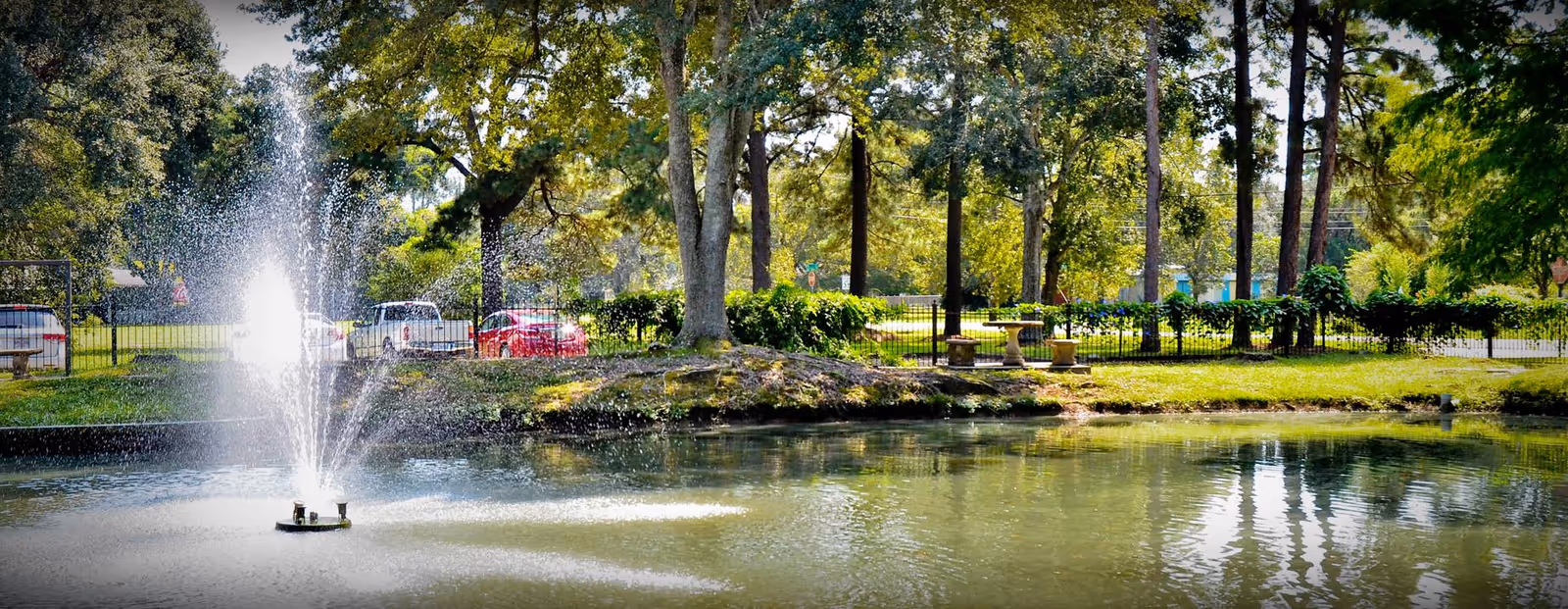 A peaceful outdoor scene featuring a pond with a water fountain spraying water upwards. Surrounding the pond are green trees and bushes, with a few parked cars visible behind a black metal fence. There is also a stone table and benches near the fence under the shade of the trees.