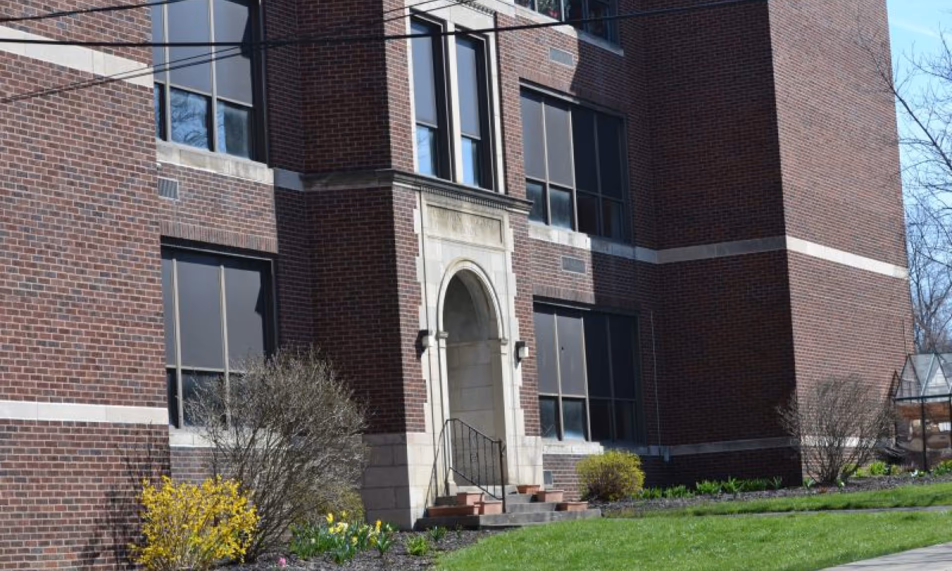 Exterior view of a brick building with large windows and an arched stone entrance. There are small bushes and flowers planted along the building, and a green lawn in the foreground under a clear blue sky.