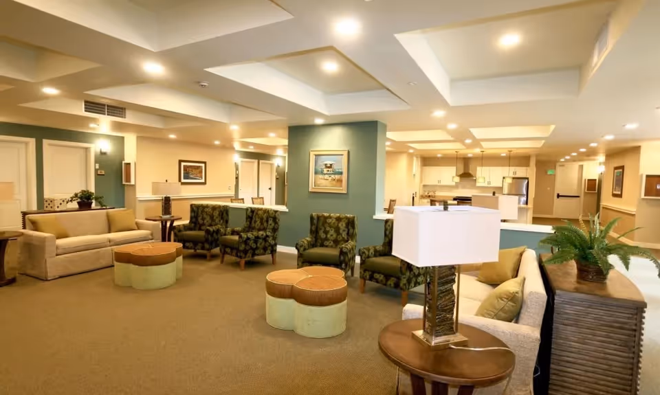 A spacious and well-lit common area in a senior living facility featuring beige sofas, patterned armchairs, round ottomans, side tables with lamps, and a green accent wall with framed artwork. The background shows a kitchen area with white cabinets and stainless steel appliances.