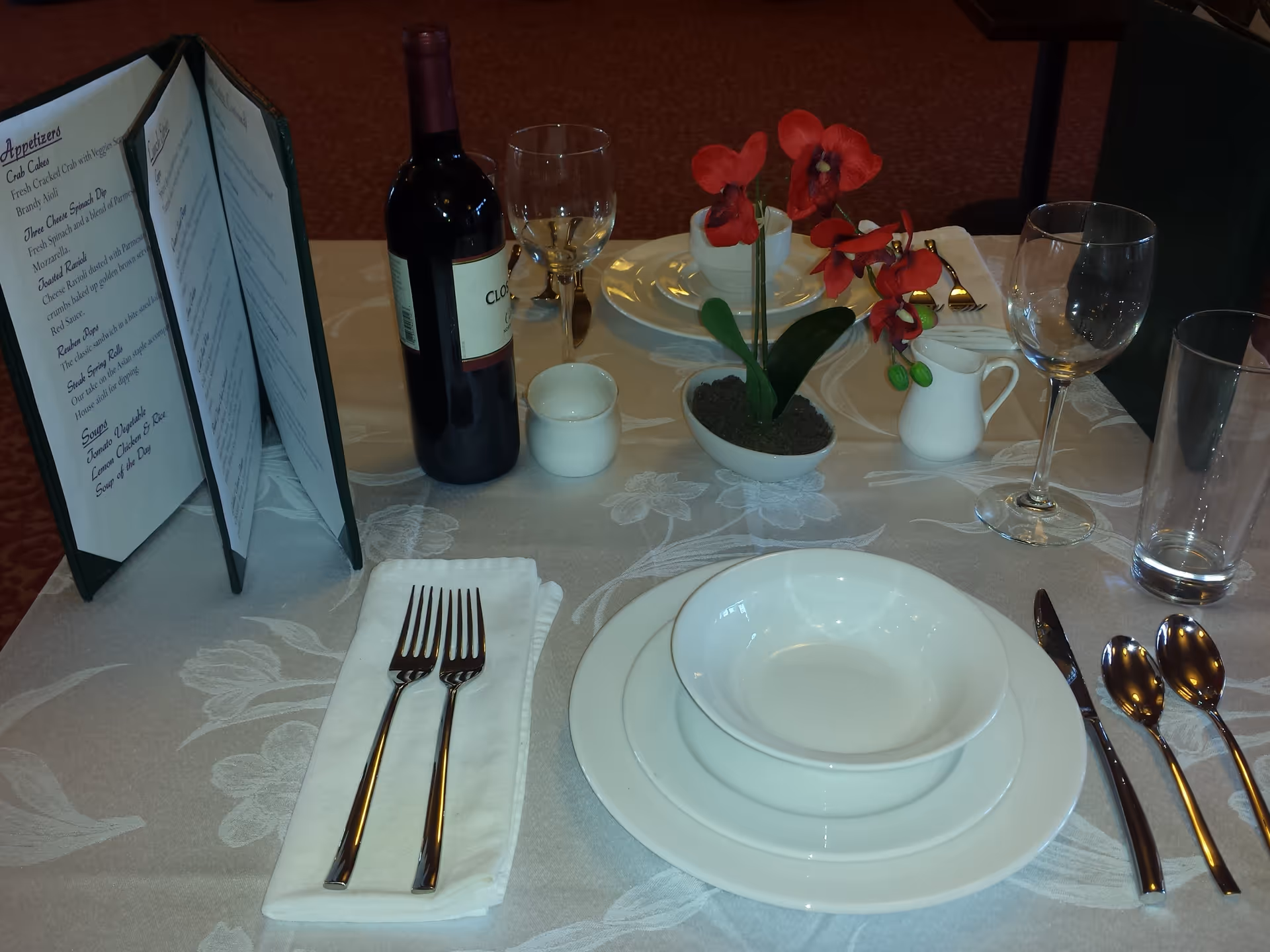 Formal dining table place setting with stacked white plates and bowl, forks on a napkin, wine bottle and glasses, a menu, and a small red orchid centerpiece.