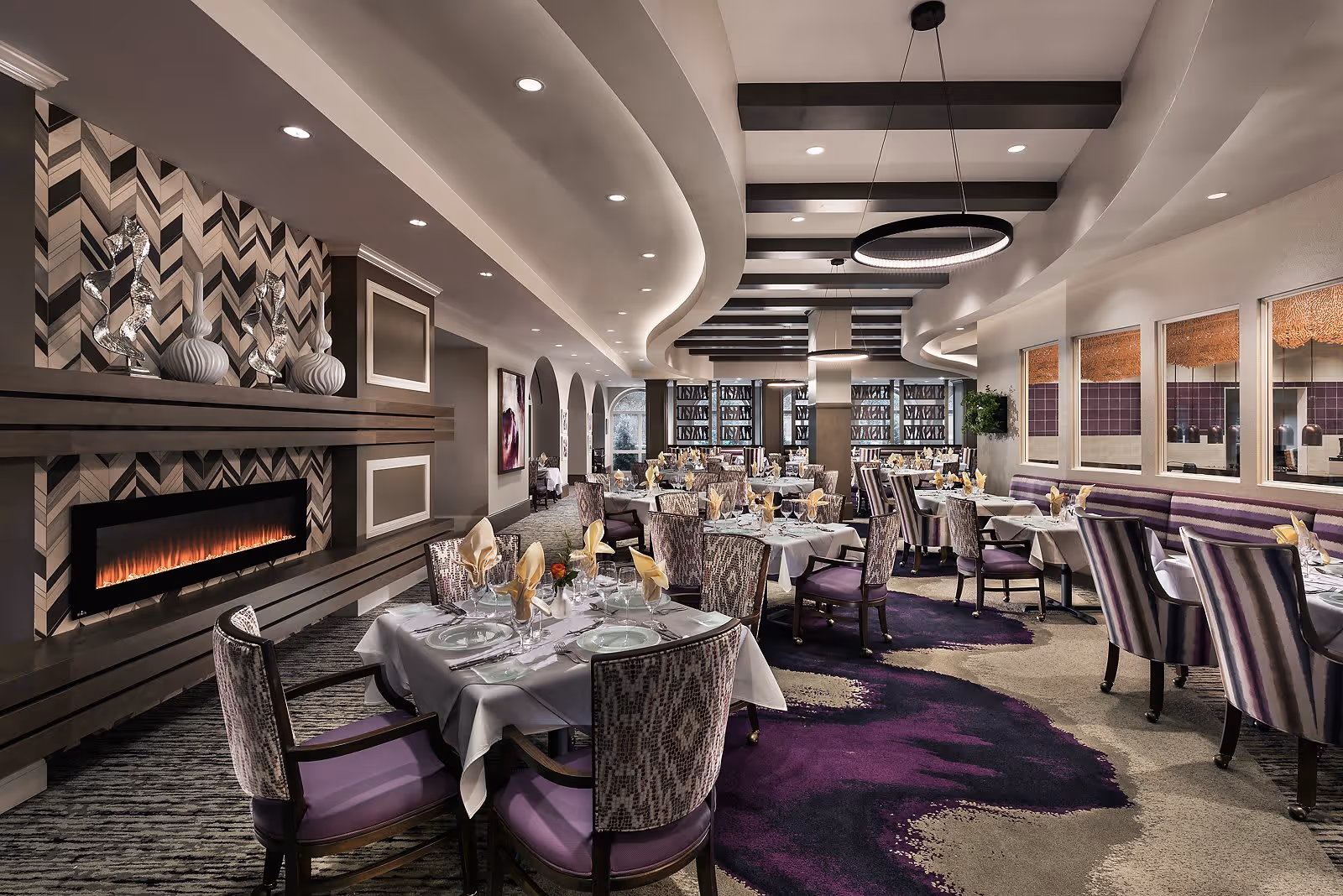 Elegant dining room with multiple tables set with white tablecloths, plates, glasses, and folded napkins. The room features patterned chairs with purple cushions, a modern fireplace with decorative vases above it, and a ceiling with dark wooden beams and circular light fixtures. The carpet has a purple and beige abstract design, and large windows with decorative panels allow natural light into the space.