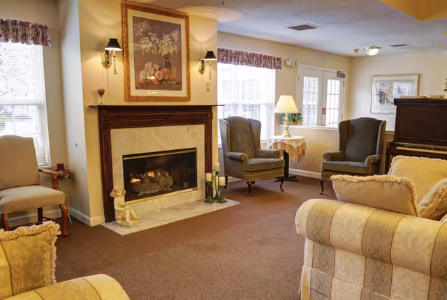 A cozy living room area with a lit fireplace framed by a wooden mantel and marble surround. Above the fireplace hangs a floral painting flanked by two wall sconces. The room features two upholstered armchairs near a window with floral valances, a side table with a lamp, and a cushioned sofa in the foreground. The carpet is brown, and the walls are painted a soft yellow. There is also a piano partially visible on the right side of the image.