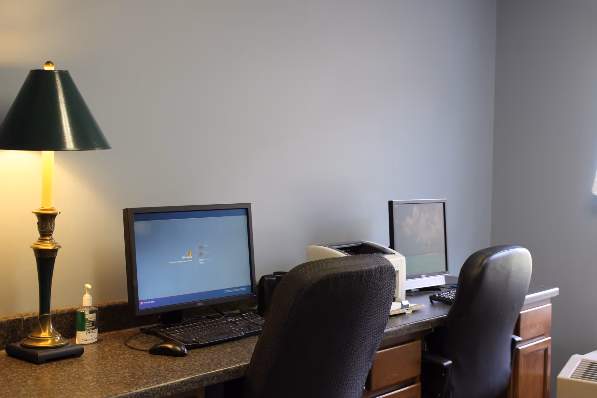 Two computer workstations with monitors and chairs on a countertop, beside a lamp and a bottle of hand sanitizer against a gray wall.
