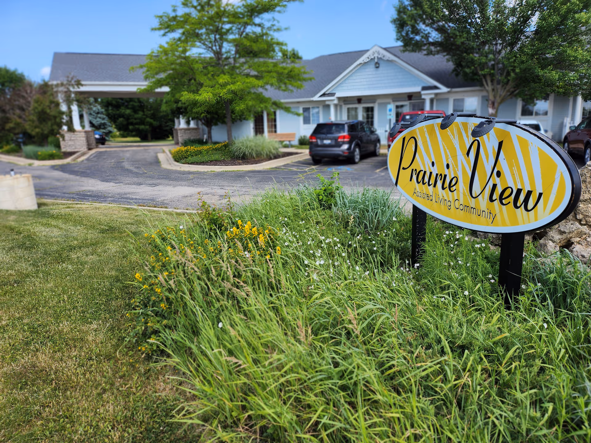View of the exterior entrance area of an assisted living community named Prairie View, showing a driveway, parked cars, a building with a covered entrance, and a garden with green grass and yellow flowers in the foreground.