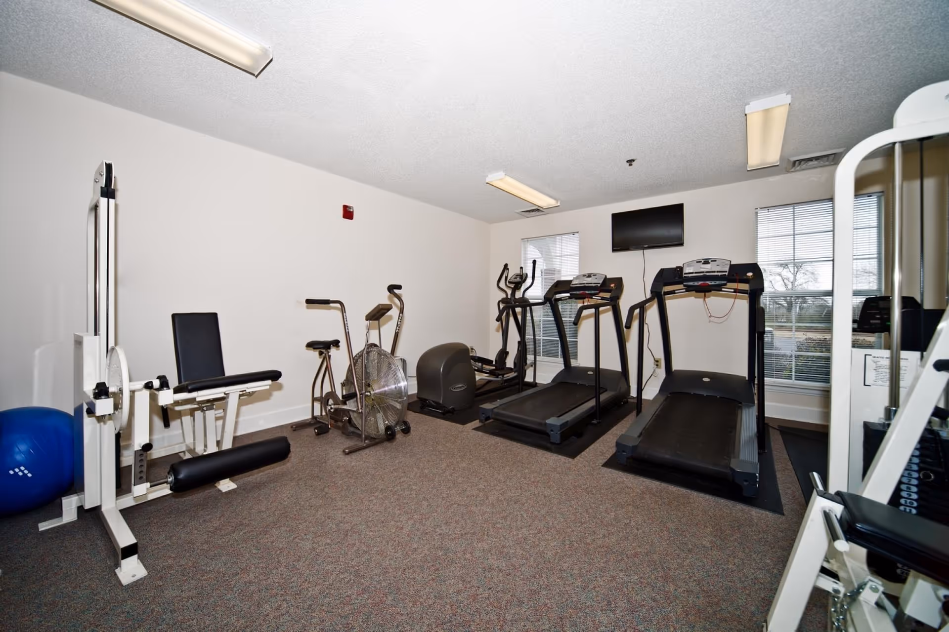 A small fitness room with exercise equipment including two treadmills, an elliptical machine, a stationary bike, a leg press machine, and a blue exercise ball. The room has carpeted flooring, white walls, two windows with blinds, and a wall-mounted TV.