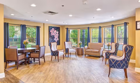 A bright and spacious living room area with large windows letting in natural light. The room features a wooden floor, a round table with four chairs on the left, and a seating area with a loveseat and four armchairs arranged around a small round table. The walls are painted yellow, and there is a large floral painting on one wall. Blue curtains frame the windows, and small flower arrangements are placed on the tables.