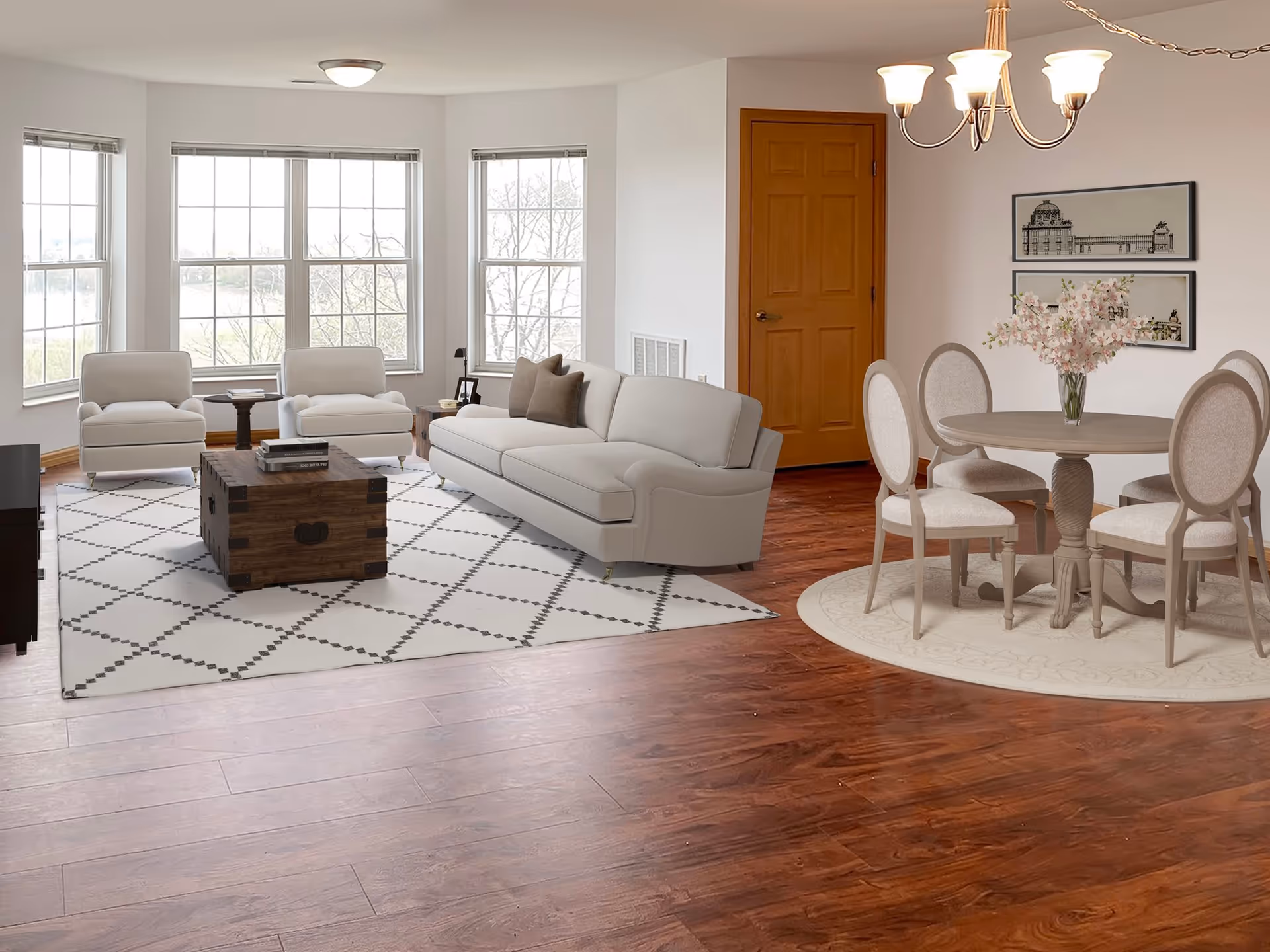 A bright living and dining area with large windows letting in natural light. The living room features two white armchairs, a white sofa with brown pillows, a wooden coffee table on a white patterned rug, and a small side table with a lamp. The dining area has a round wooden table with four upholstered chairs on a circular rug, a vase with pink flowers on the table, and two framed architectural prints on the wall. The floor is wooden, and there is a wooden door in the background.