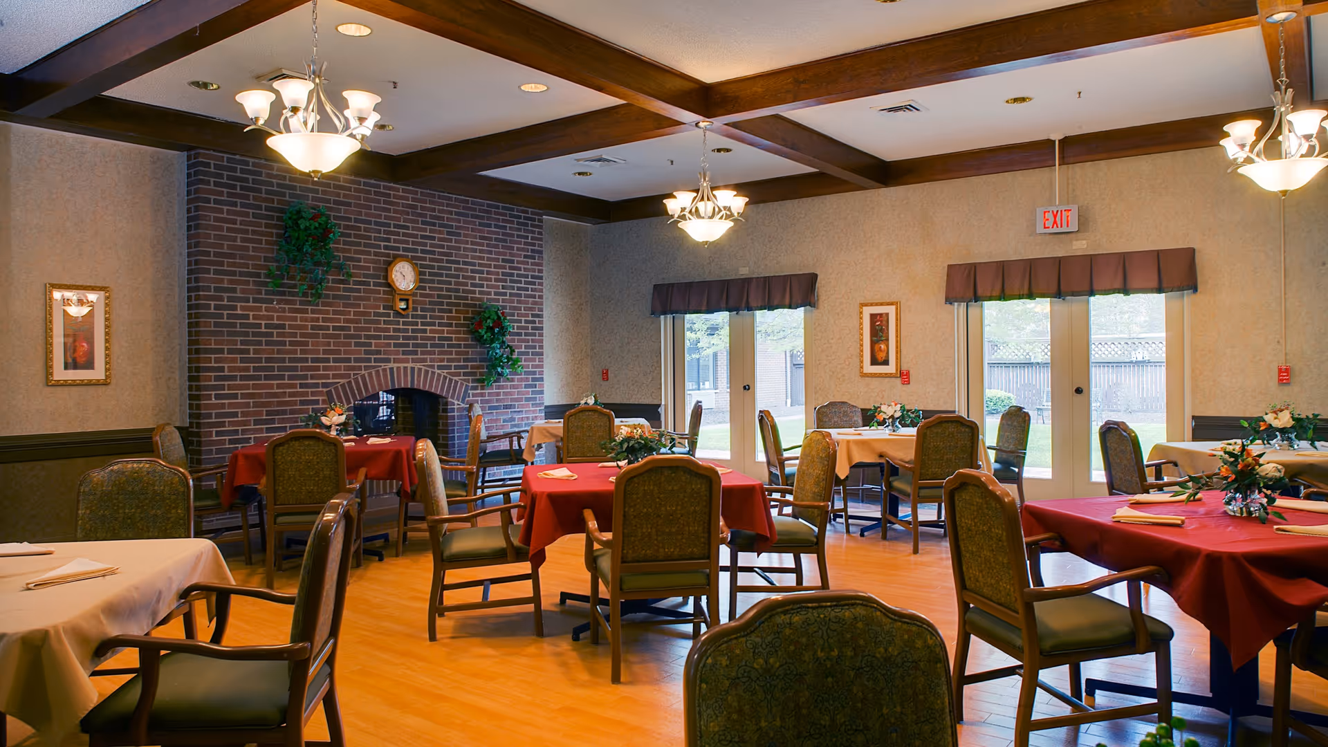 A dining room with multiple tables covered in red and beige tablecloths, each set with napkins and floral centerpieces. The room features wooden chairs with green upholstery, a brick fireplace adorned with plants and a clock, beige walls with framed artwork, and large windows with valances letting in natural light. Ceiling lights hang from wooden beams.