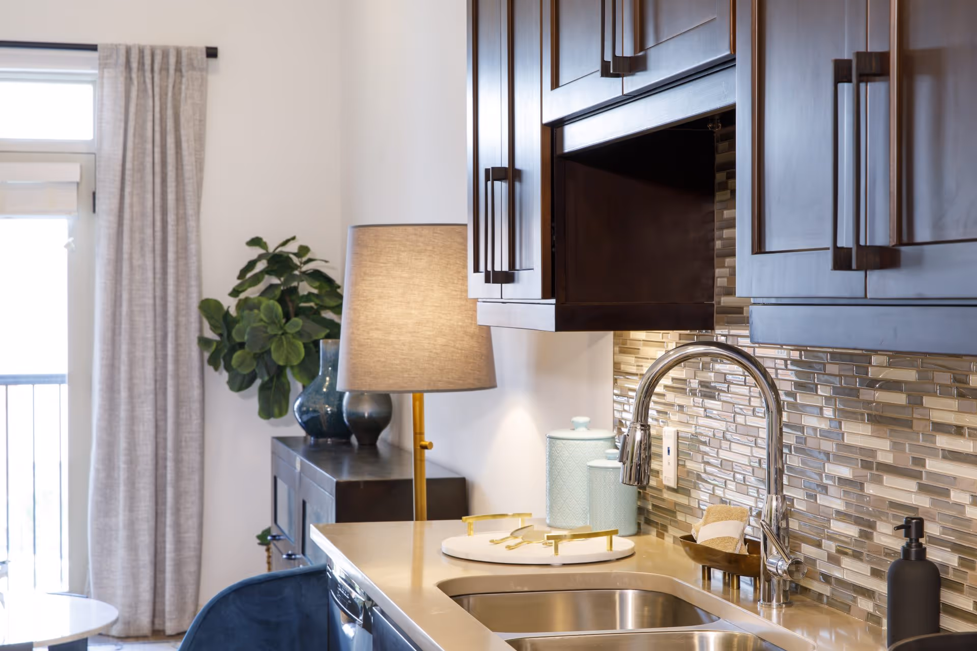 A modern kitchen area with a stainless steel sink and chrome faucet, beige countertop, and dark wooden cabinets. The backsplash features a mosaic tile design in neutral tones. On the counter, there are two light blue canisters, a soap dispenser, and a small tray with rolled towels. In the background, there is a lamp with a beige shade, a dark sideboard with decorative vases, and a window with light gray curtains.