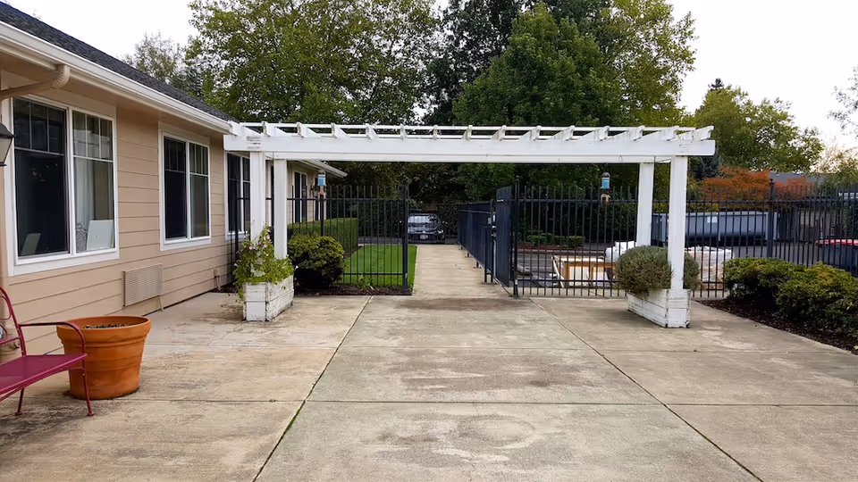 Paved courtyard with a white pergola, planters, and a metal fence beside a beige building.