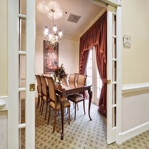 A formal dining room with a long wooden table surrounded by eight upholstered chairs. The room features a chandelier hanging from the ceiling, a framed artwork on the wall, and large windows covered with heavy red curtains. The floor is carpeted with a patterned design, and the room is viewed through white double doors with glass panes.