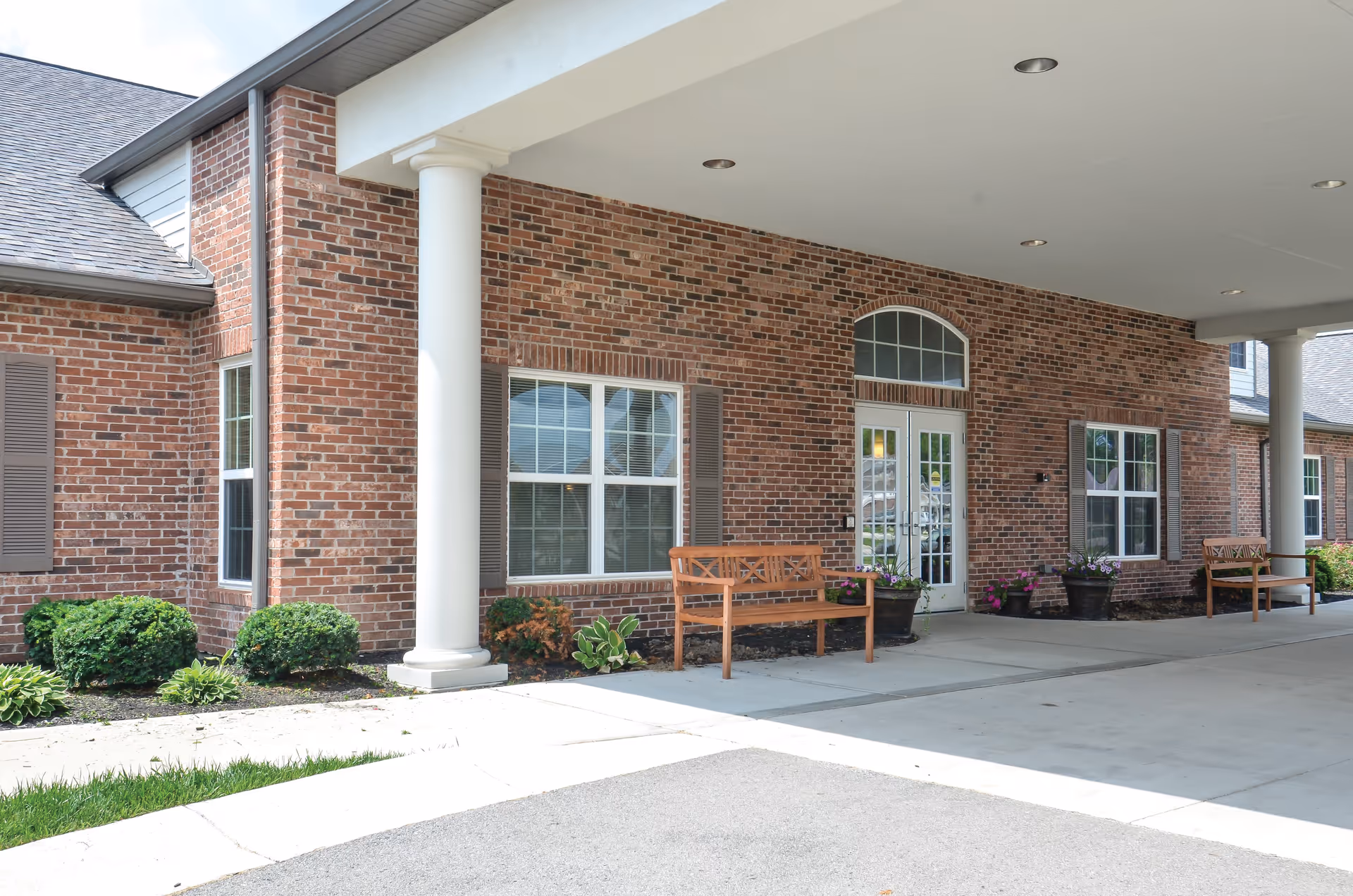 Entrance area of a brick building with white columns supporting a covered driveway. There are two wooden benches and several potted plants near the windows and door. The building has large windows with shutters and a glass door with an arched window above it.