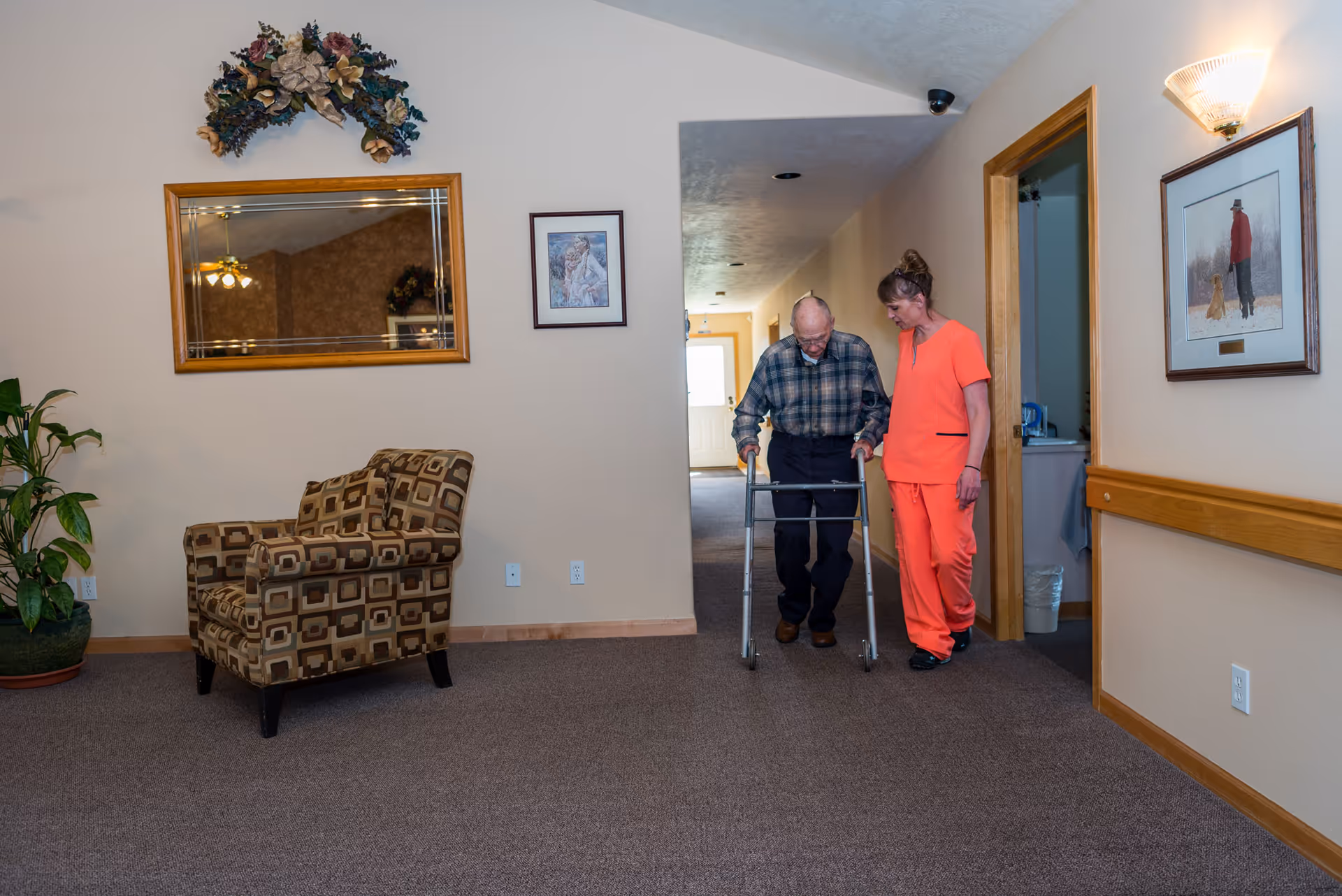 An elderly man using a walker is assisted by a caregiver dressed in orange scrubs as they walk down a carpeted hallway in an assisted living facility. The hallway features a patterned armchair, a potted plant, a large mirror with floral decoration above it, and framed artwork on the walls.