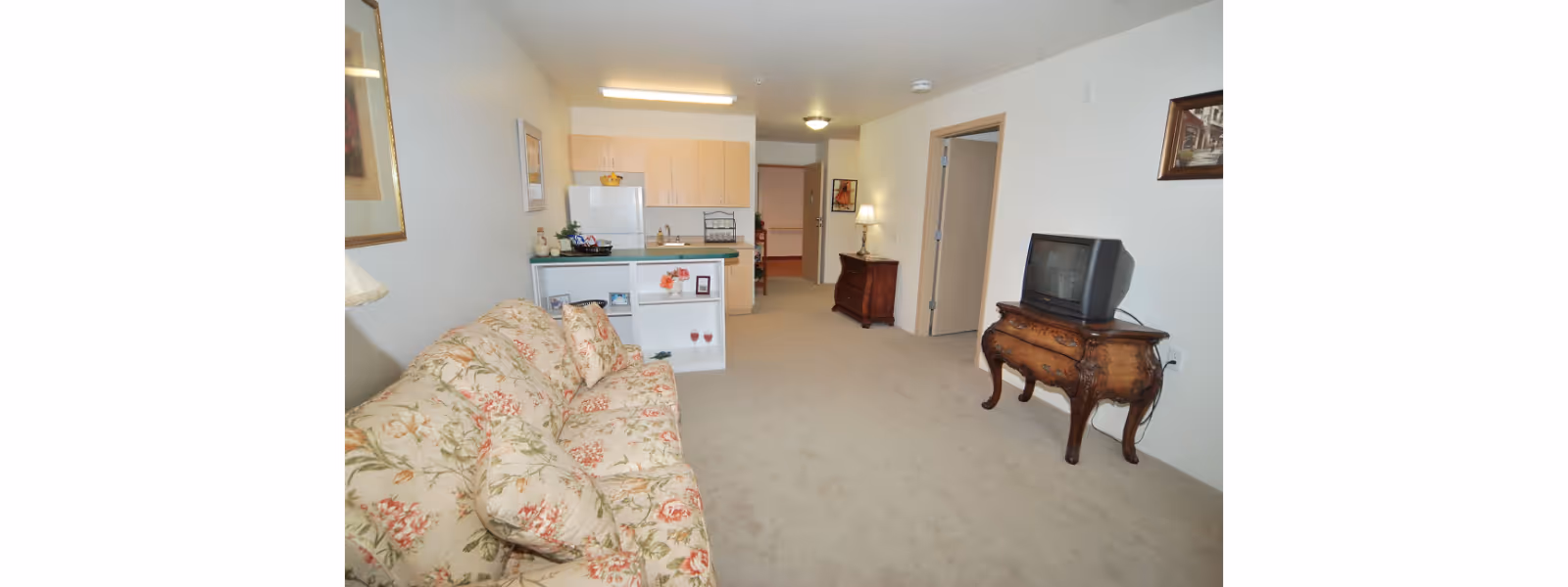Interior view of a senior living facility apartment showing a floral patterned sofa on the left, a small wooden table with an old-style TV on the right, and a kitchenette with light wood cabinets and a white refrigerator in the background. The room has beige carpeting and white walls with framed artwork. A hallway leads to other rooms in the back.