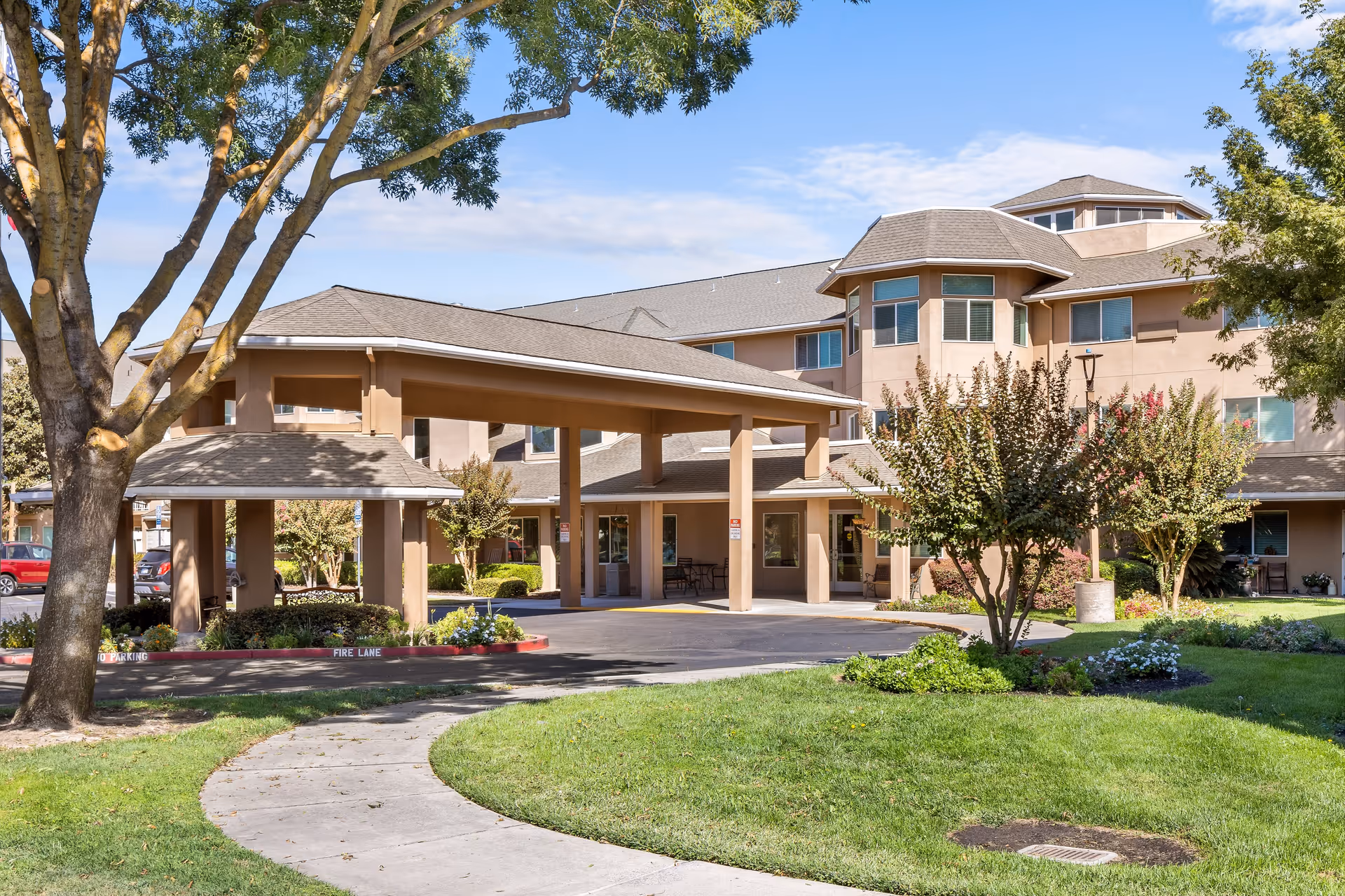 Exterior view of The Hampshire - A Provincial Senior Living Community building with a covered entrance, surrounded by trees, shrubs, and a well-maintained lawn under a partly cloudy sky.