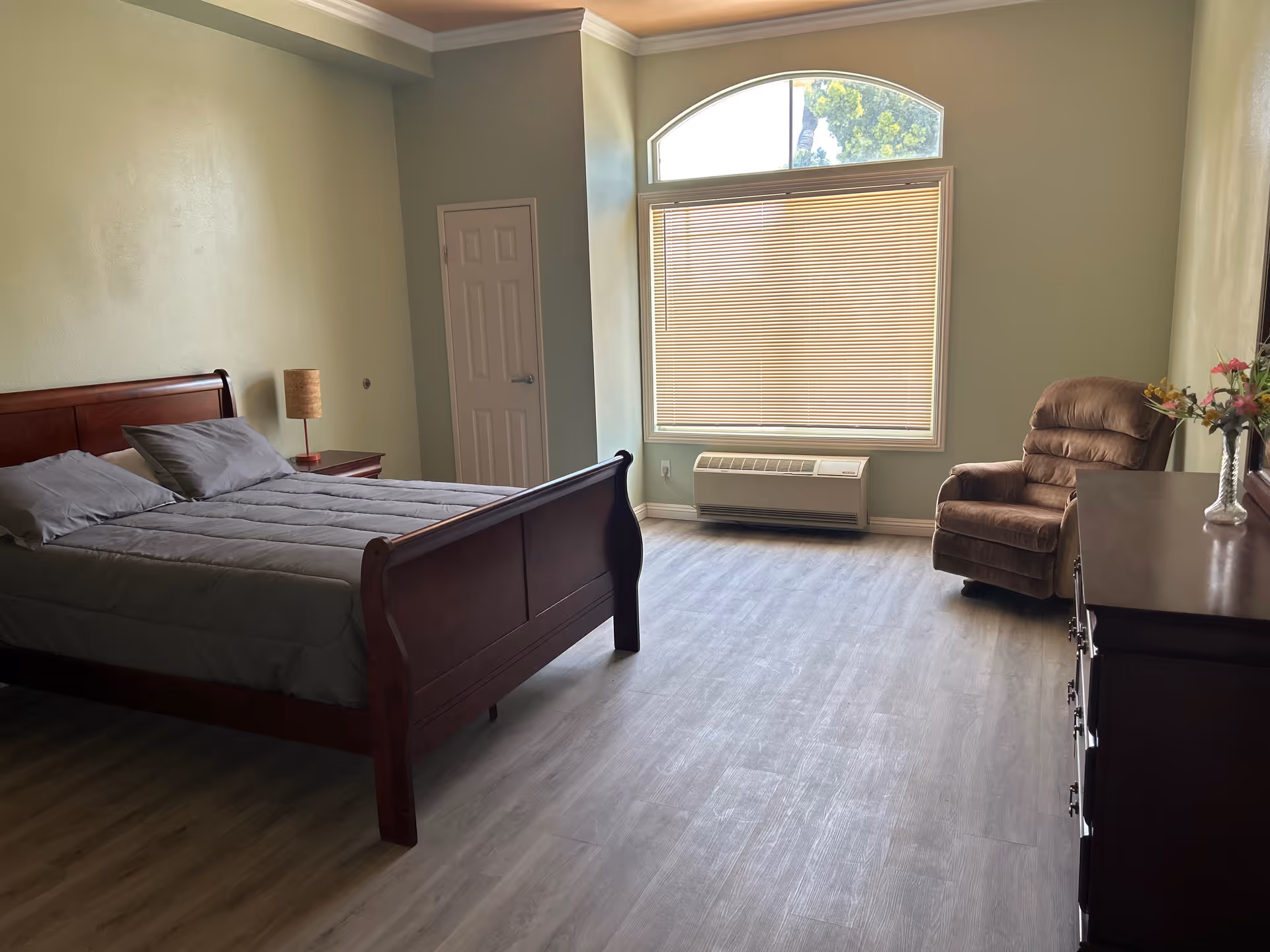 A spacious bedroom with a wooden bed frame and gray bedding, a matching wooden nightstand with a lamp, a large window with blinds and an arched top, a brown recliner chair, and a wooden dresser with a vase of flowers on top. The room has light-colored walls and wood flooring.