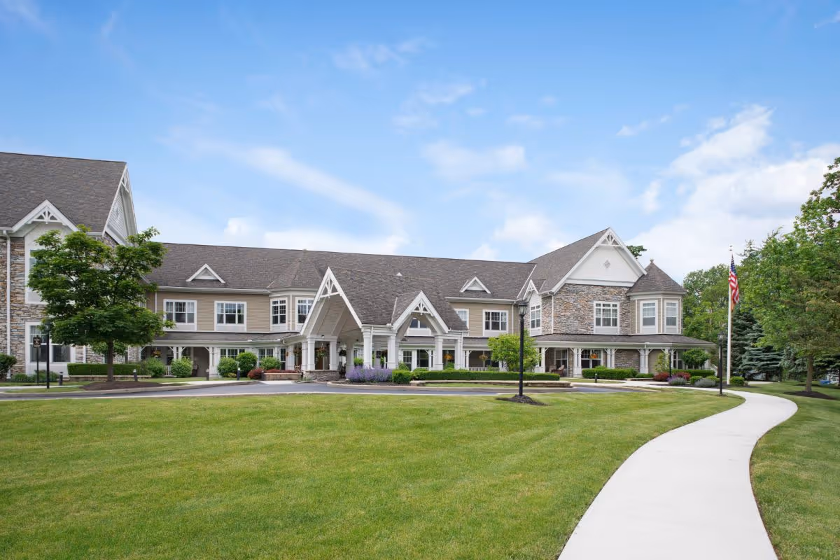 Exterior view of a large, two-story senior living facility building with stone and beige siding, a covered entrance, well-maintained green lawn, trees, and a curved sidewalk leading to the entrance under a partly cloudy blue sky.