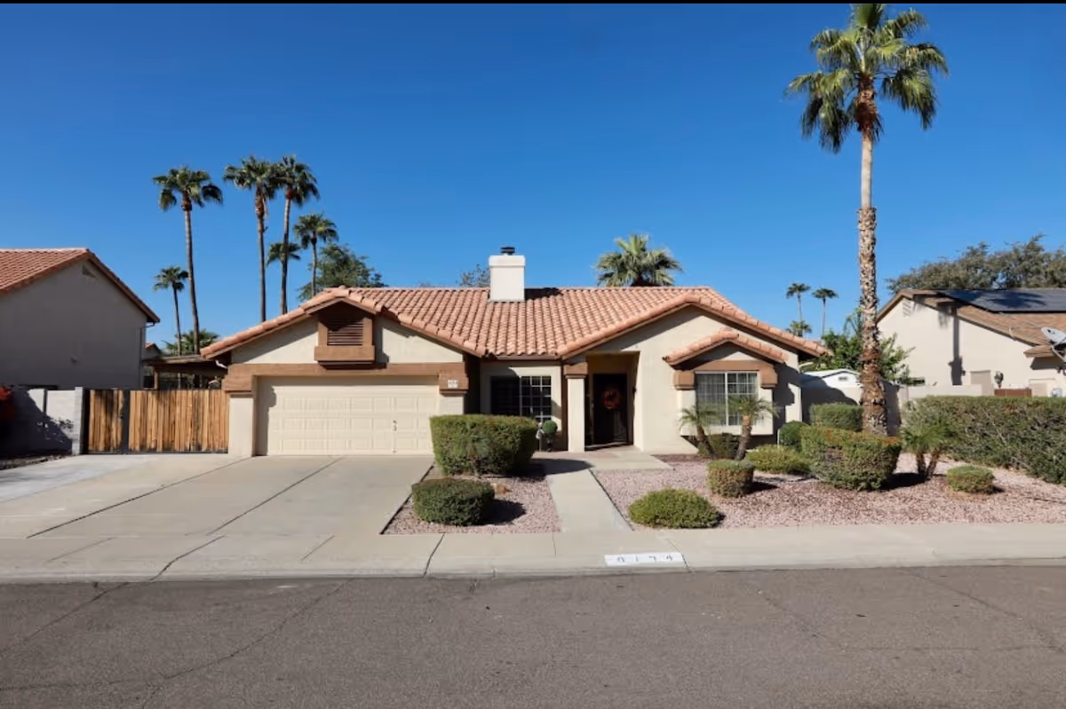 Single-story stucco house with a tiled roof, attached garage, desert landscaping and palm trees under a clear blue sky.