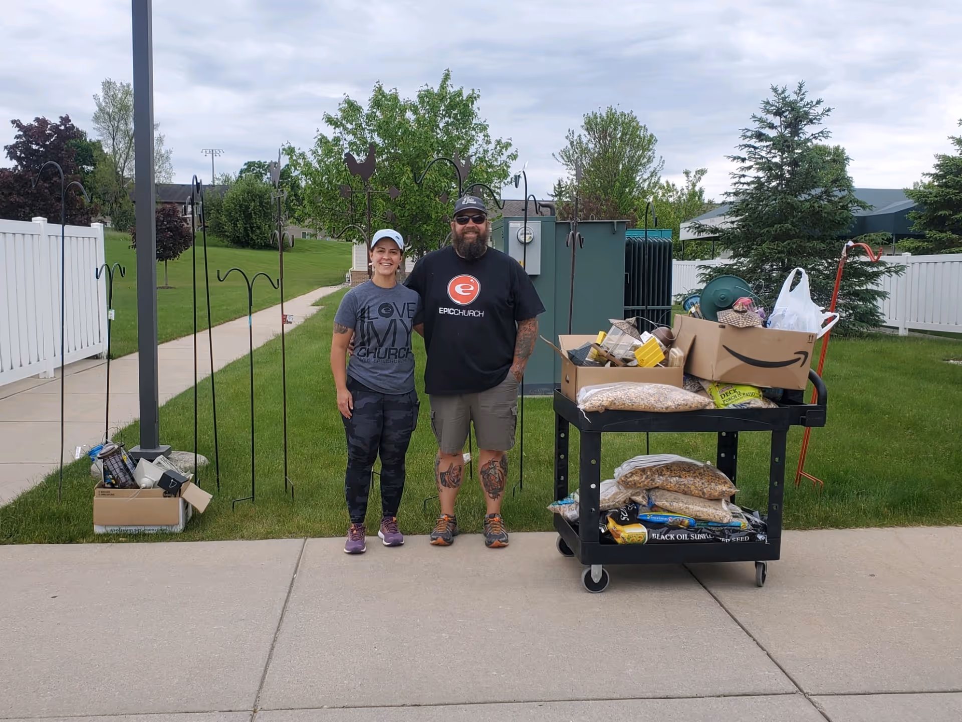 Two people standing outside on a sidewalk next to a black cart filled with boxes and bags of various items. They are in a grassy area with trees, a white fence, and some utility equipment in the background. The person on the left is wearing a gray t-shirt, camo pants, purple shoes, and a white cap. The person on the right is wearing a black t-shirt, shorts, sunglasses, a black cap, and has tattoos on their legs.