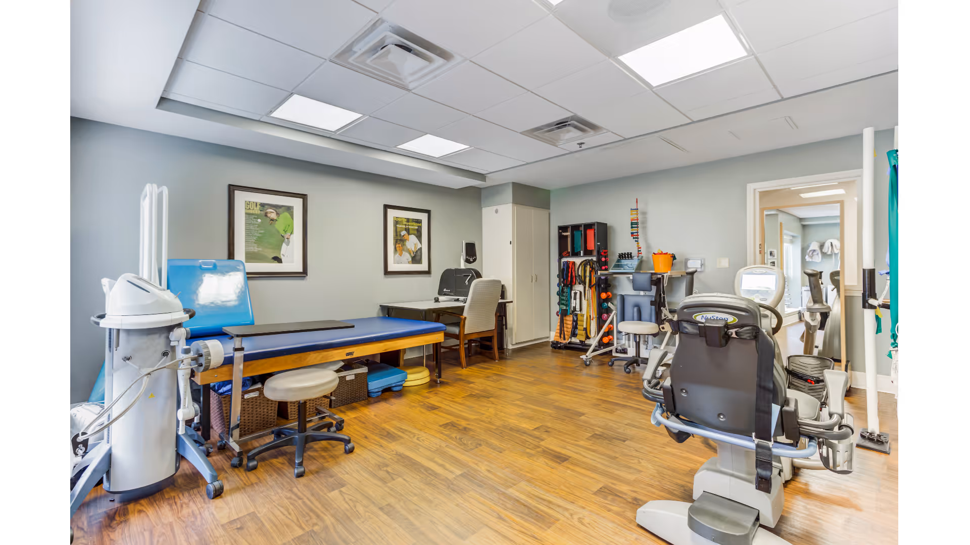 Well-lit physical therapy room with exercise machines, a treatment table, equipment storage, and wood flooring.