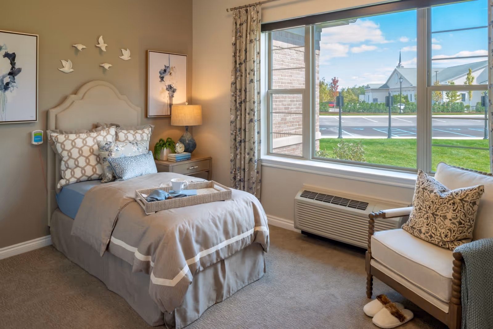 A cozy bedroom with a single bed dressed in beige and patterned pillows, a bedside table with a lamp, books, and a small plant. There are two framed floral artworks and decorative white bird sculptures on the wall above the bed. A large window shows a view of a parking lot and a building outside. An armchair with a patterned pillow and a green throw blanket sits near the window, with a pair of slippers on the floor beside it.