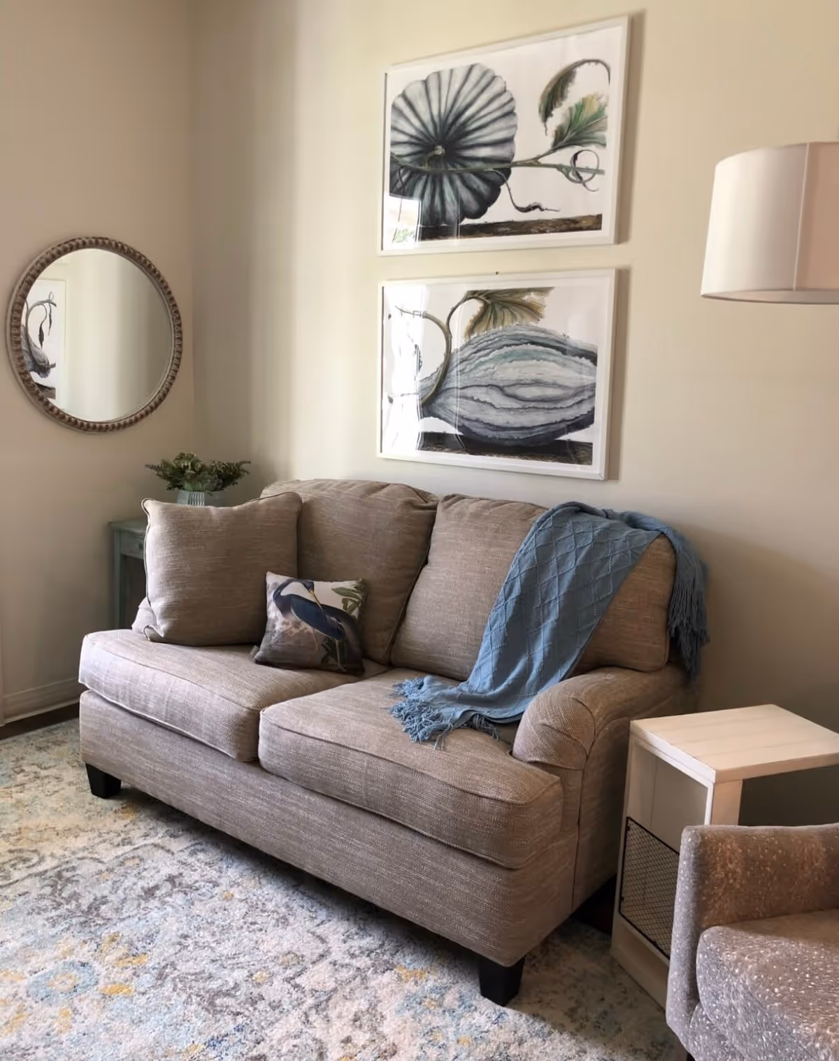 A cozy living room area with a beige sofa adorned with a blue throw blanket and a decorative pillow featuring a bird. Above the sofa are two framed botanical prints. To the left, there is a round mirror on the wall and a small green side table with a plant. A white side table and a textured armchair are partially visible on the right. The floor has a patterned rug with soft colors.