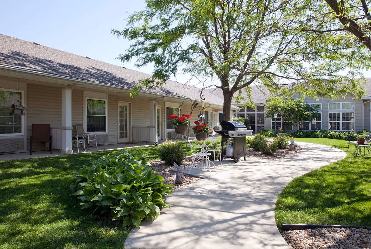 Sunny courtyard with a curved concrete walkway, lawn and shrubs, patio chairs, potted flowers and a grill in front of single-story building units.