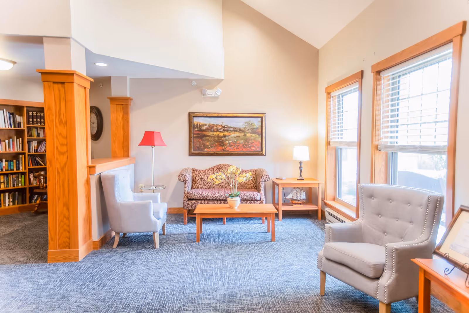 Bright sitting area with armchairs, a patterned loveseat, coffee table, lamps, bookshelves, and large windows.