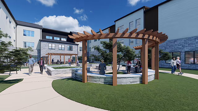 Outdoor courtyard area at a senior living facility with a circular stone seating area topped by a wooden pergola. Several people are walking and sitting around the courtyard, surrounded by multi-story buildings under a blue sky with some clouds.