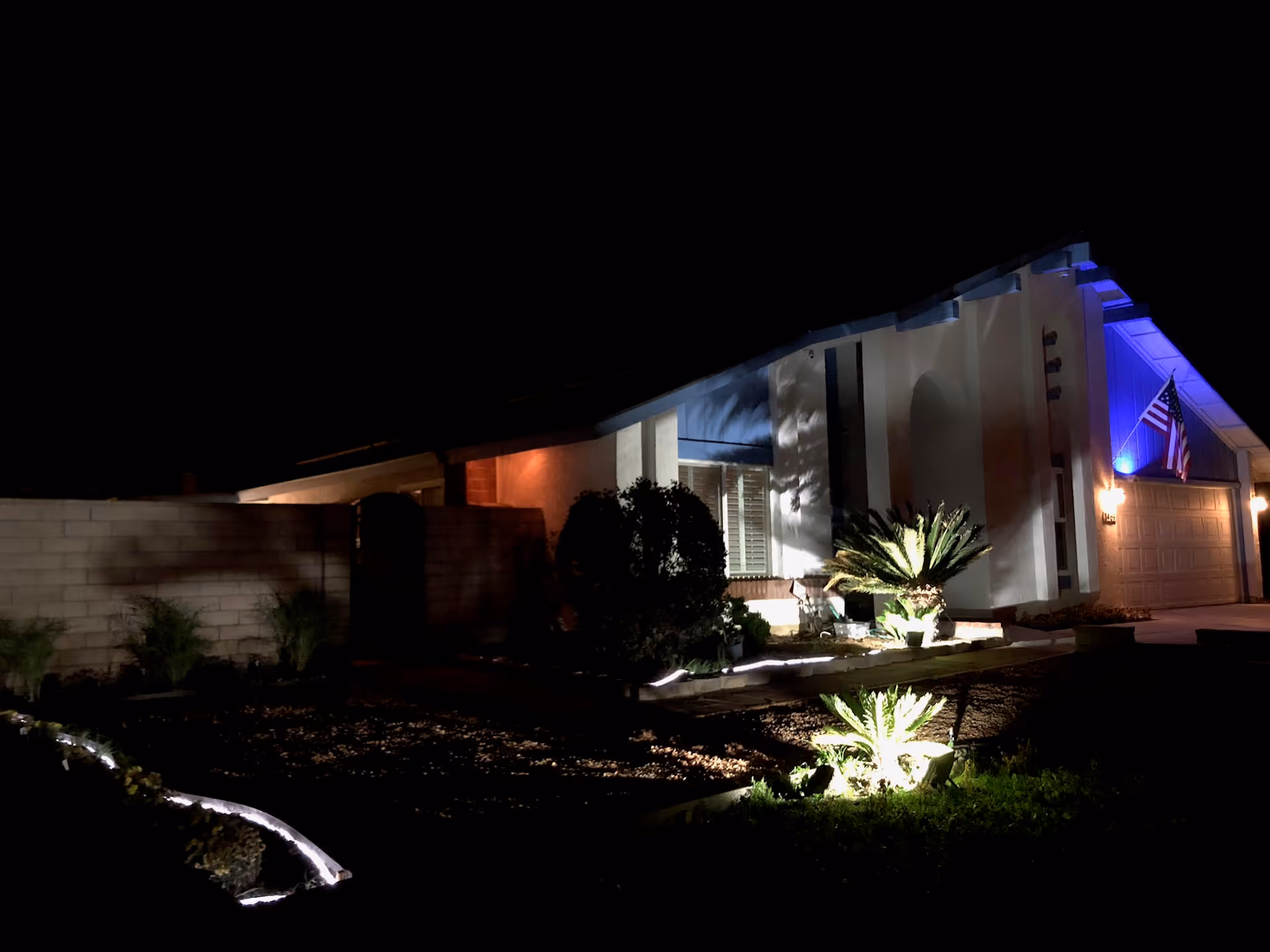 Night view of a residential house with exterior lighting illuminating the front yard plants and the American flag mounted near the garage door.