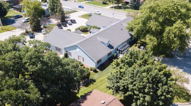 Aerial view of a single-story building surrounded by trees and greenery, with a parking lot and road visible in the background. The building has a gray roof and light-colored exterior walls with red shutters on the windows.