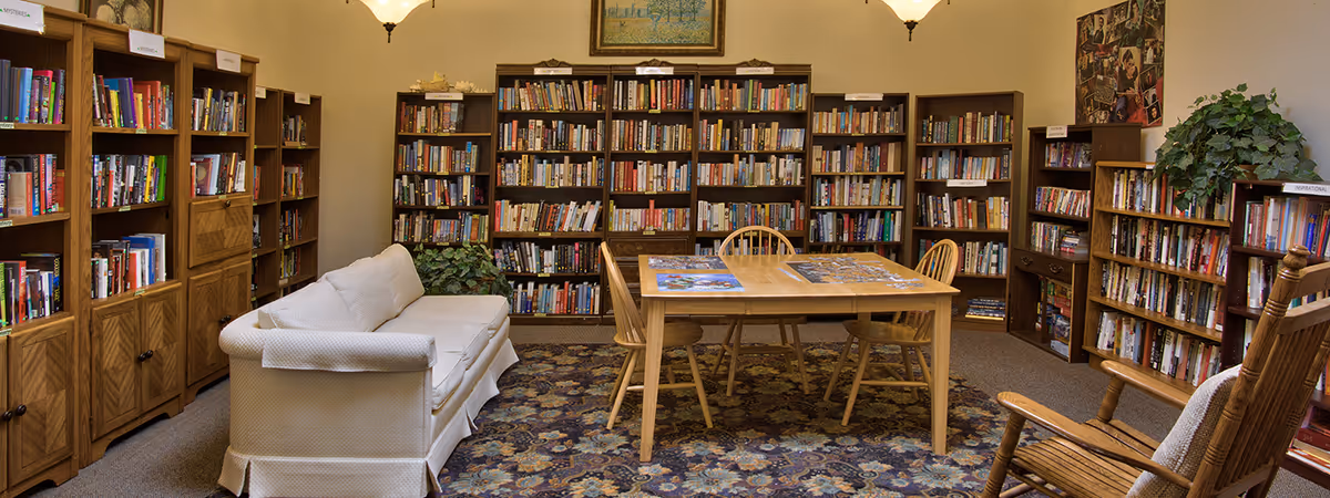 Cozy library-style common room with bookshelves lining the walls, a couch, a wooden table with chairs and a rocking chair.