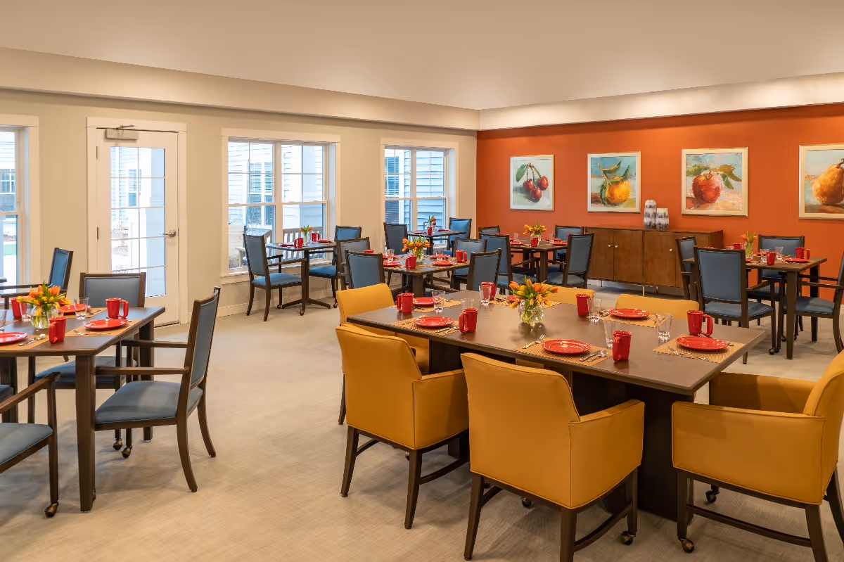 A bright dining room with multiple tables set for a meal. The tables have red plates, red mugs, and glassware, with small floral centerpieces. The room features large windows letting in natural light, yellow and blue chairs, and a warm orange accent wall adorned with framed fruit paintings.