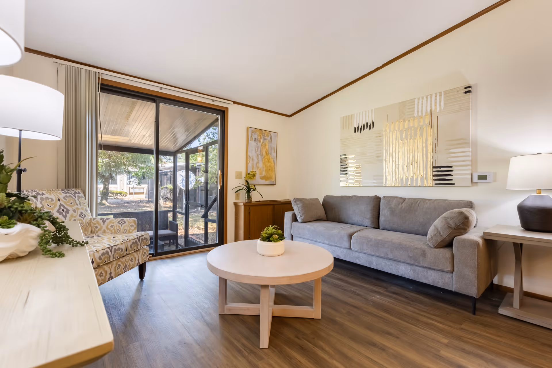 A cozy living room with a gray sofa, a patterned armchair, a round wooden coffee table with a small plant, and a sliding glass door leading to an outdoor patio. The room has wooden flooring, light-colored walls, and modern artwork above the sofa. There are two lamps providing warm lighting.