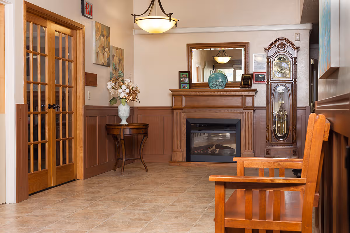 A cozy interior hallway featuring a wooden bench on the right, a wooden grandfather clock, and a wooden fireplace mantel with a large mirror above it. There is a small round table with a vase of flowers on the left side, and double wooden doors with glass panes. The floor is tiled, and the walls are painted beige with wooden wainscoting. A ceiling light fixture hangs above.
