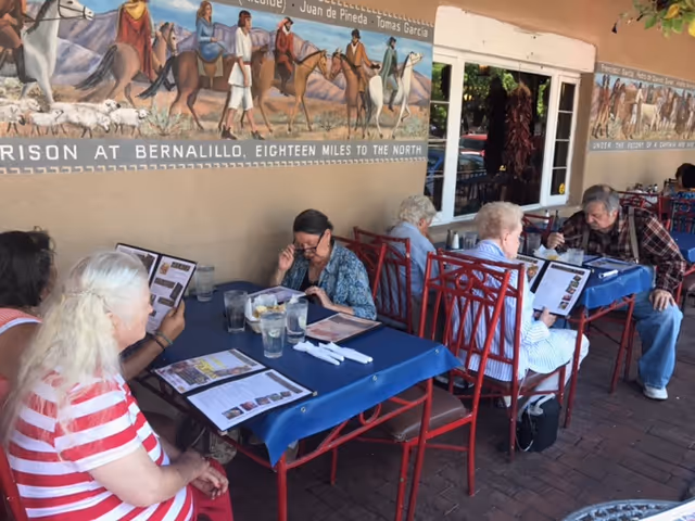 Several elderly people sitting at outdoor tables with blue tablecloths, reading menus and preparing to order food. The setting appears to be a patio area with a mural on the wall depicting people on horseback and sheep in a rural scene.