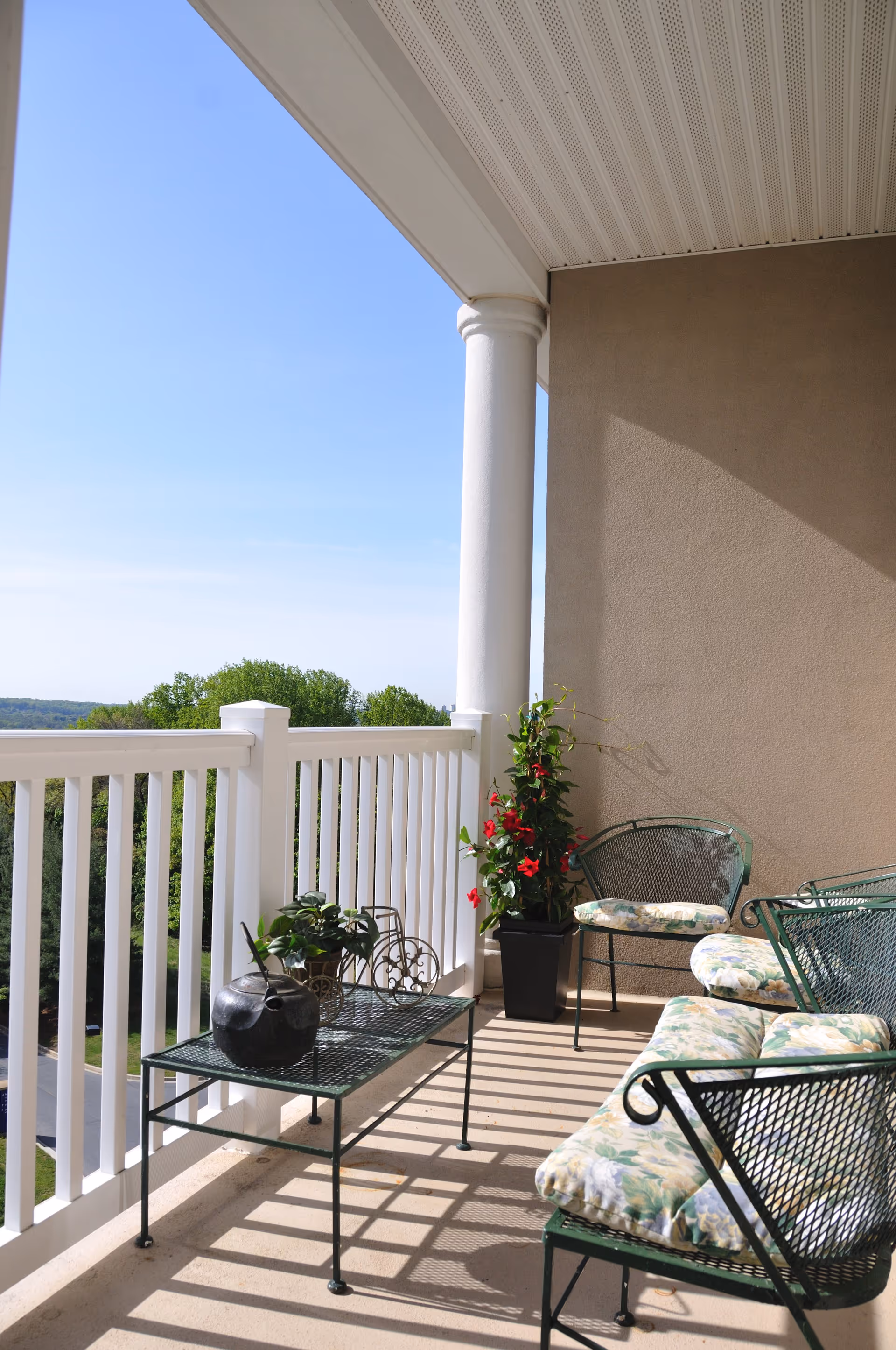 A sunny outdoor balcony with white railing and a beige wall. The balcony has green metal furniture with floral cushions, including chairs and a table. There are potted plants on the table and in a black planter, with red flowers climbing up a trellis. The sky is clear and blue with trees visible in the background.
