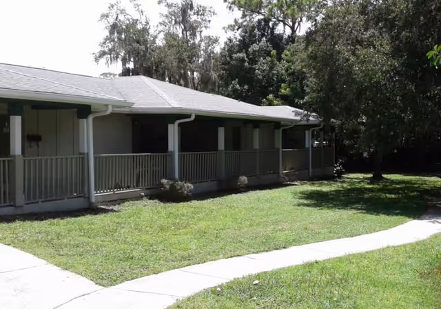 Single-story building with a covered porch and railing, surrounded by green grass and trees under a clear sky.