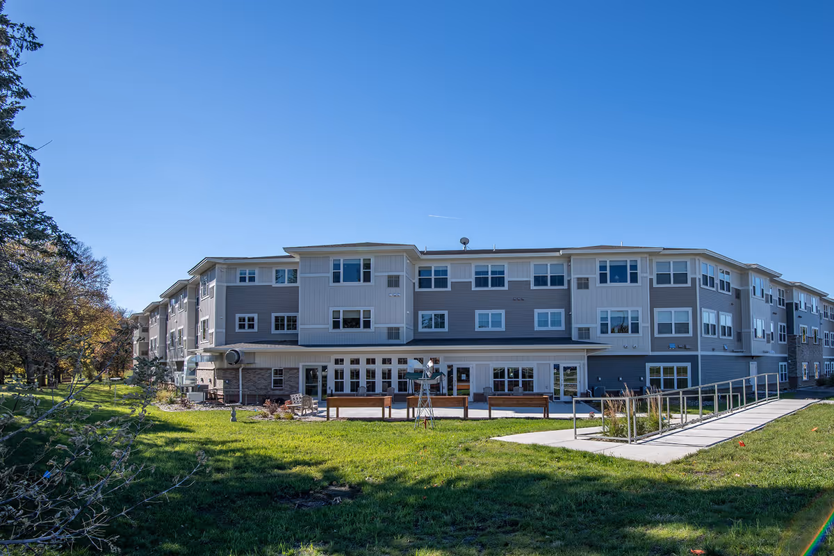 Exterior view of a three-story senior living facility building with multiple windows, a patio area with benches, a wheelchair accessible ramp, and green lawn surrounded by trees under a clear blue sky.