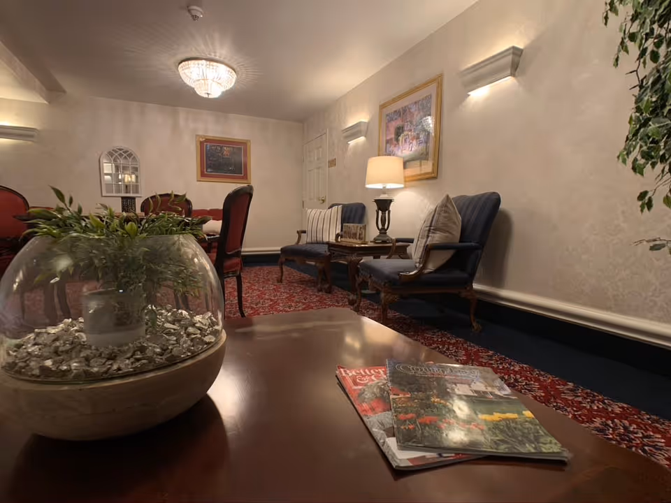 A cozy sitting area in an assisted living facility with two upholstered armchairs, a small wooden table with a lamp, framed artwork on the walls, a decorative plant in a glass bowl on a wooden coffee table, and magazines placed on the table. The room has patterned carpet and soft lighting from a ceiling chandelier and wall sconces.