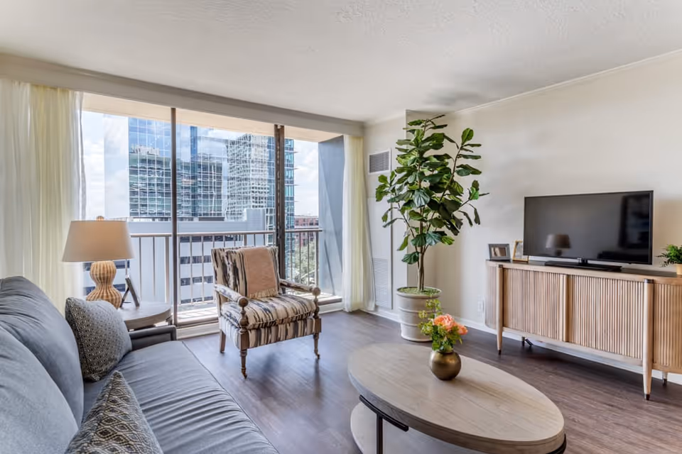 A bright living room with a large window and sliding glass door leading to a balcony overlooking tall buildings. The room features a gray sofa with patterned pillows, a wooden armchair with a cushion, a round wooden coffee table with a small vase of flowers, a wooden TV console with a flat-screen TV, a large potted plant, and a table lamp on a side table.