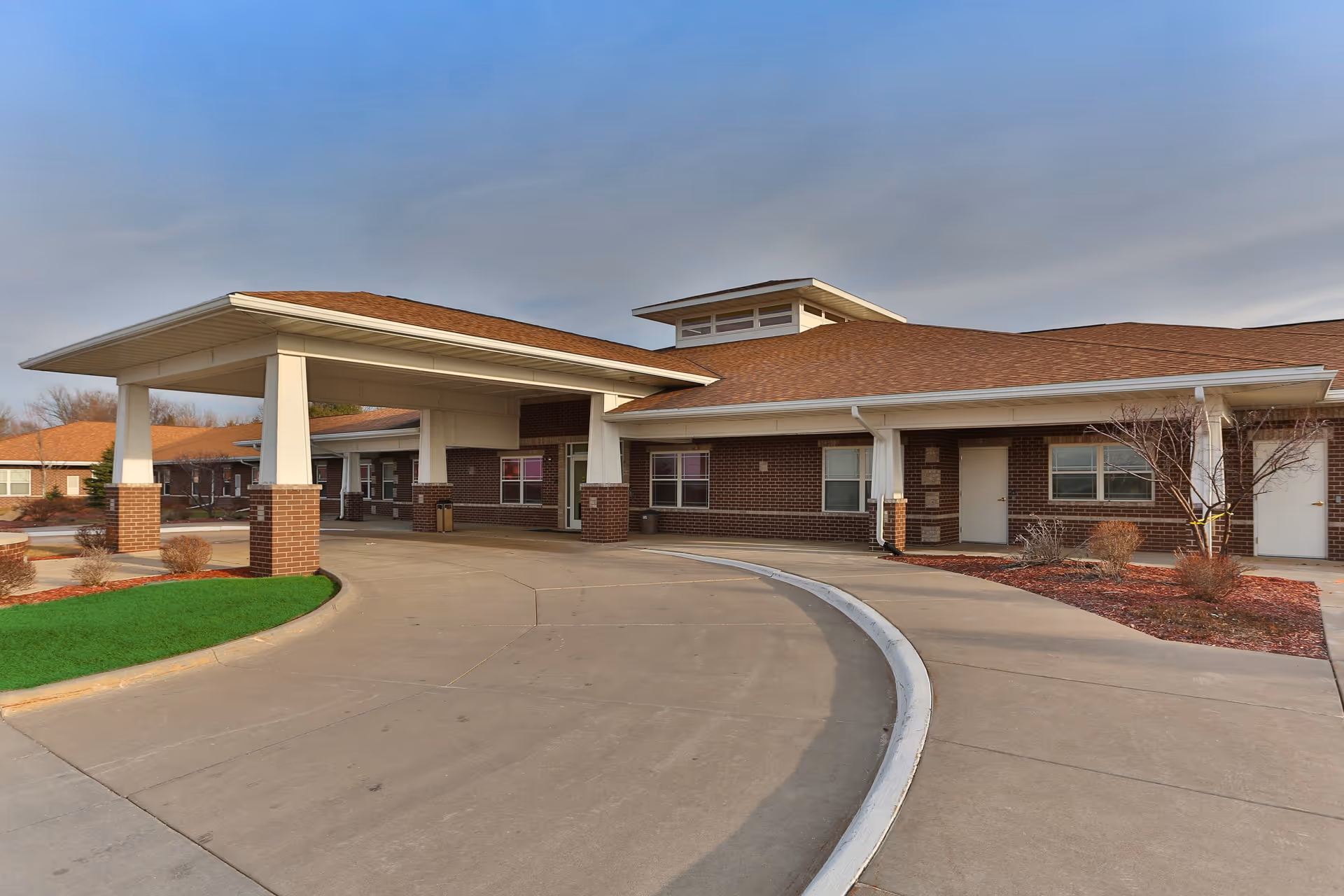 Exterior view of a senior living facility building with a covered entrance driveway, brick walls, and a brown shingled roof under a clear sky.