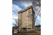 Tall multi-story senior residence building with a parking area, trees, and a partly cloudy sky.
