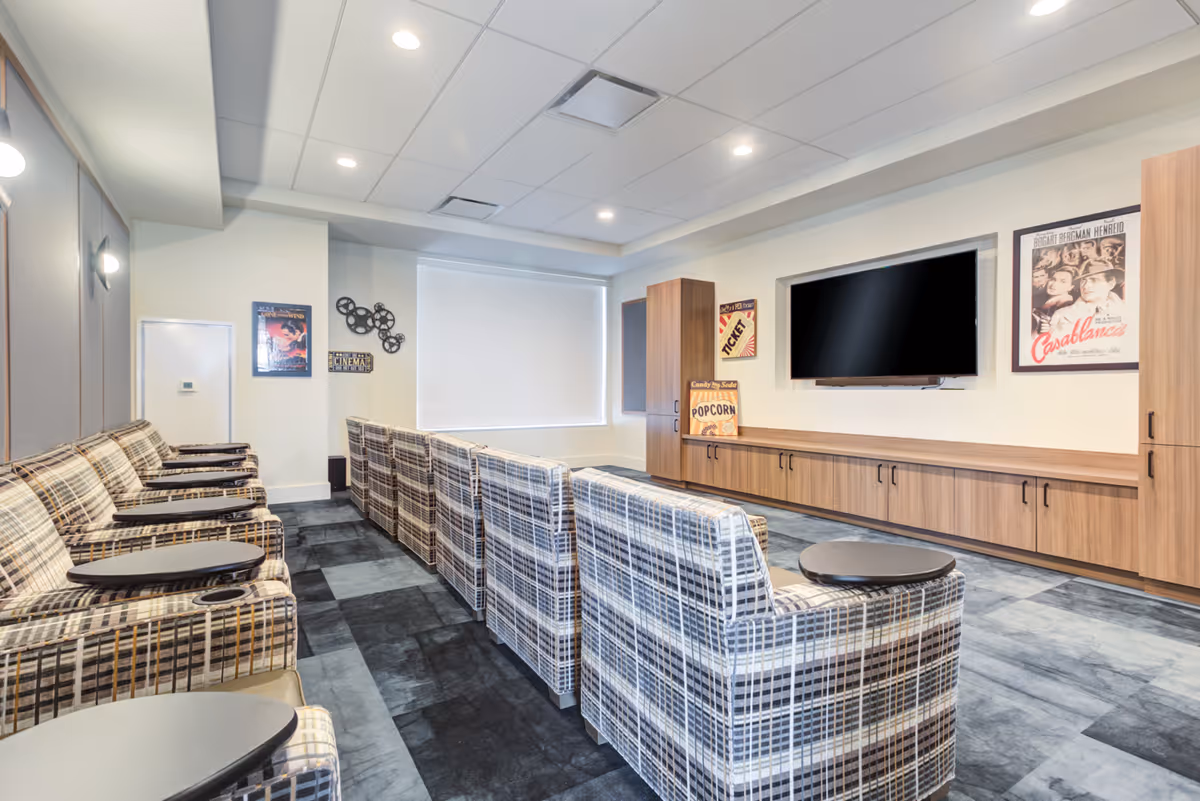 A modern media room with rows of plaid upholstered chairs equipped with small black tables and cup holders, facing a large flat-screen TV mounted on a wooden cabinet. The room has light-colored walls decorated with vintage movie posters and a film reel wall decoration, with a window covered by a white blind.