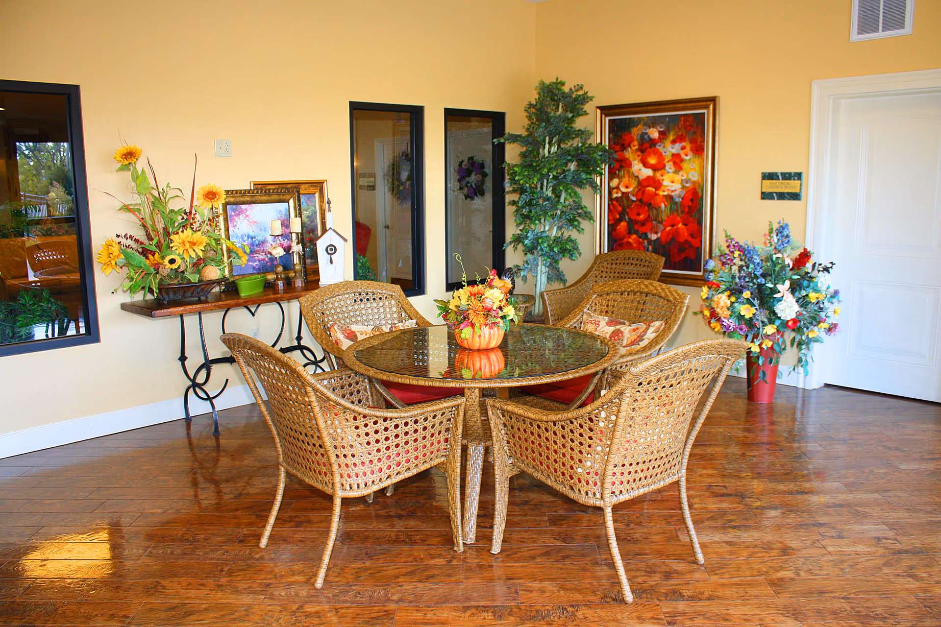 A cozy seating area with a round glass-top table surrounded by five wicker chairs with red cushions. The room has wooden flooring and yellow walls adorned with colorful floral paintings and several flower arrangements. There is a small table against the wall with decorative items including framed pictures, candles, and a birdhouse. A tall green plant is placed near the corner.