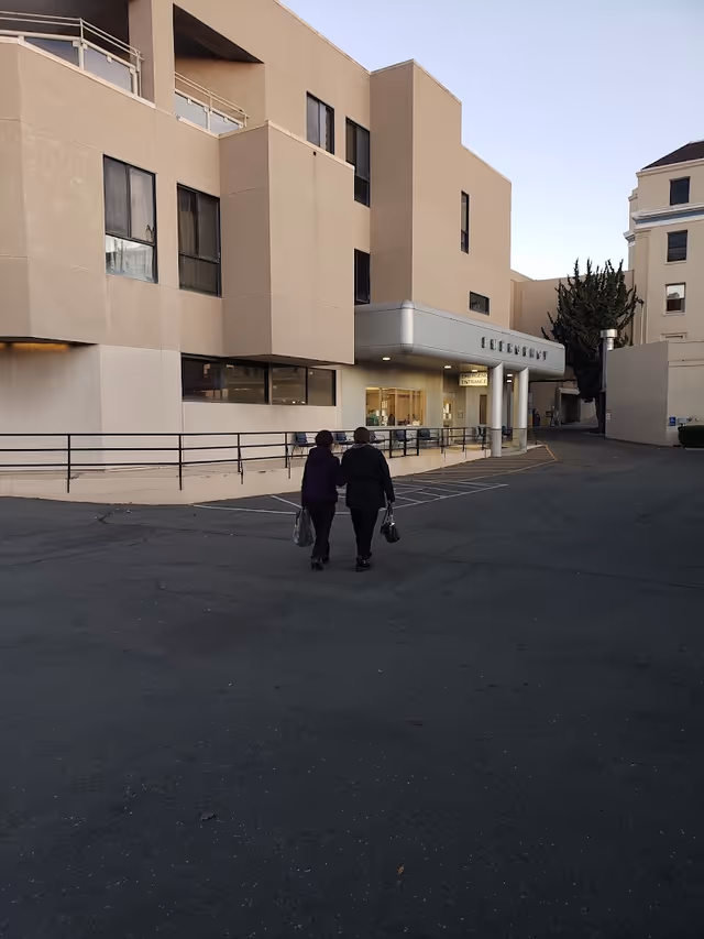 Two people walking towards the entrance of a beige multi-story hospital building with a sign above the entrance that reads 'EMERGENCY'.