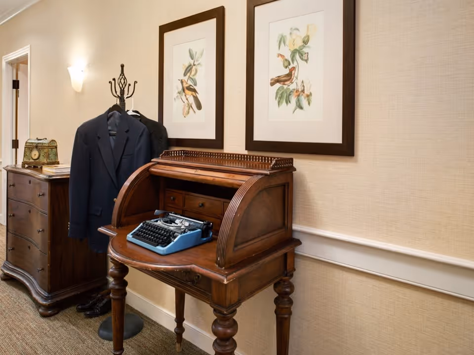 A vintage wooden roll-top desk with a blue typewriter on it, positioned against a beige textured wall. Above the desk are two framed botanical prints featuring birds and flowers. To the left of the desk is a wooden dresser with a decorative box and some books on top. A coat rack with a dark blazer hanging on it stands between the dresser and the desk. The floor is carpeted and a doorway is visible in the background.