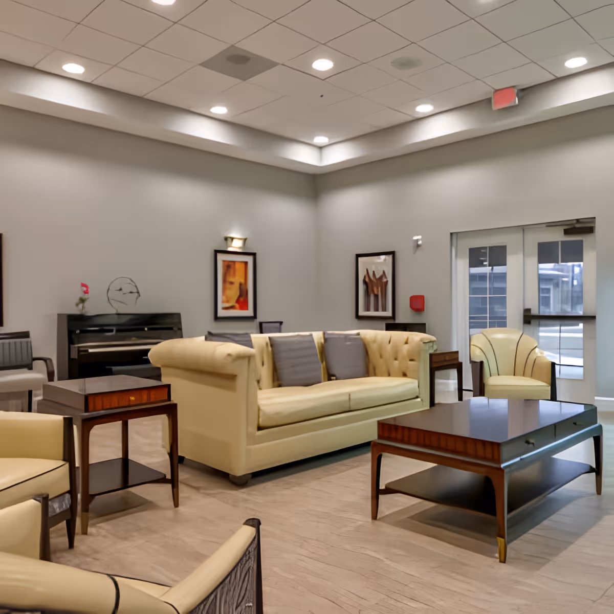 A well-lit living room area with beige sofas and armchairs arranged around a wooden coffee table. The room has light-colored walls with framed artwork, a piano in the corner, and double glass doors leading outside.