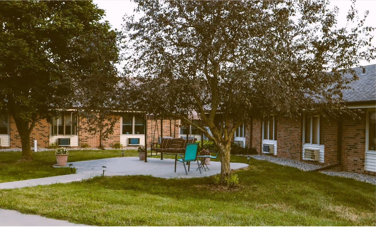 A grassy courtyard with a concrete patio, chairs and a swing beneath trees in front of a single-story brick building.