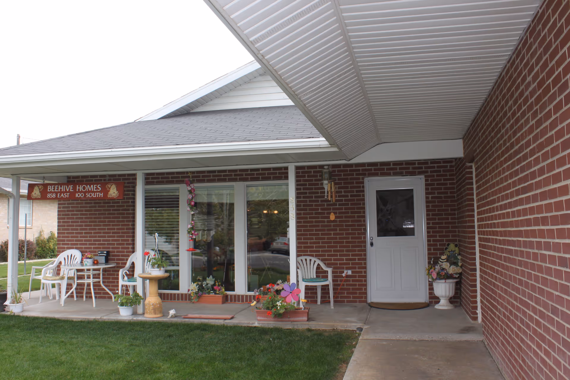 Covered front porch area of a brick building with a white door and large window. The porch has white plastic chairs, a small table, various potted plants, and decorative items including a wind chime and flower pinwheel. A sign reading 'BEEHIVE HOMES 858 EAST 100 SOUTH' hangs above the porch.